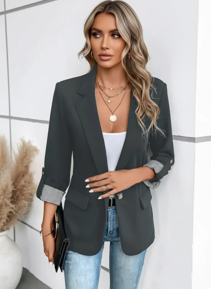 A woman wearing a black casual slim-fit cropped suit jacket with a white top and blue jeans. Accessories include a necklace, a watch, and a handbag.