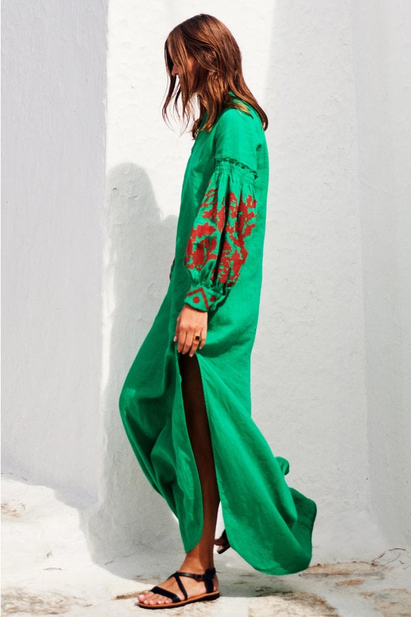 Woman wearing a green dress with red embroidery against a white wall.