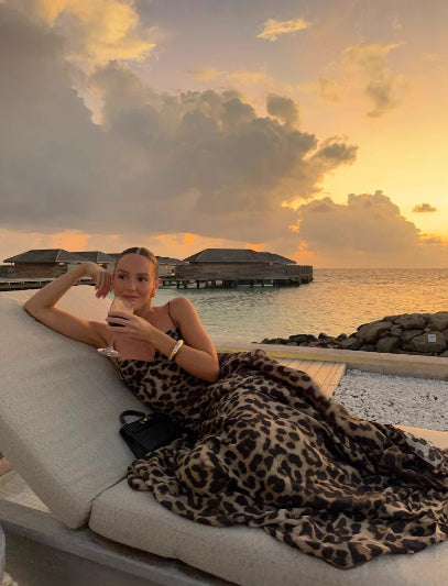 Woman in leopard print dress lounging on a sunbed by the ocean at sunset.