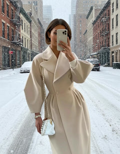 Person wearing a beige coat taking a mirror selfie in front of a building.