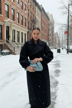 A person wearing a long black coat with a large lapel and lantern sleeves, holding a blue bag and a smartphone.
