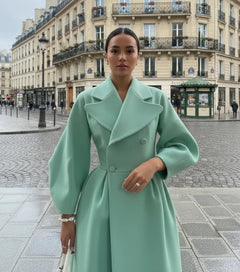 A person wearing a long beige coat with a large lapel and lantern sleeves, holding a blue bag and a smartphone.