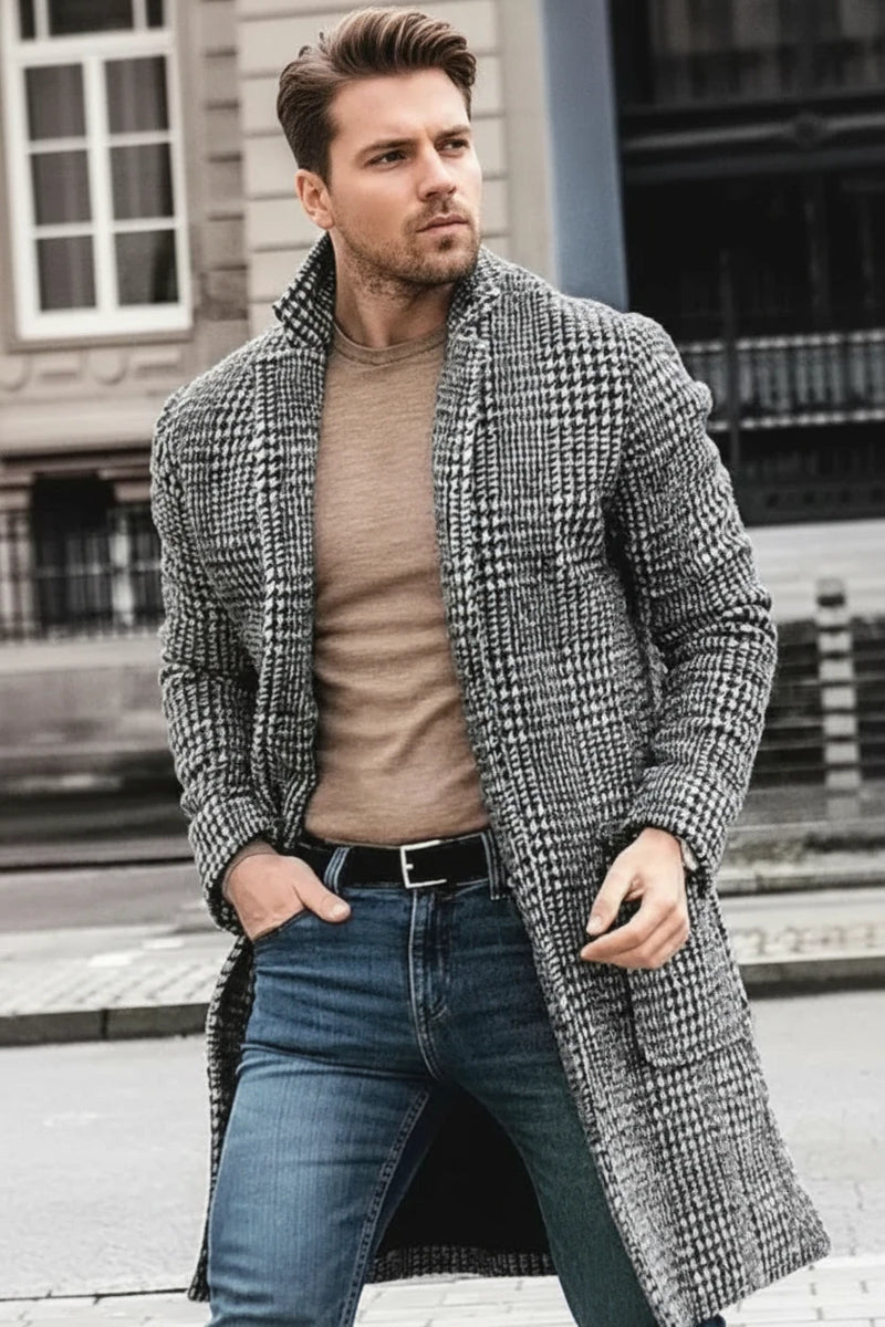 A man wearing a mid-length checkered woolen coat in gray, with a suit collar, over a beige sweater and blue jeans.