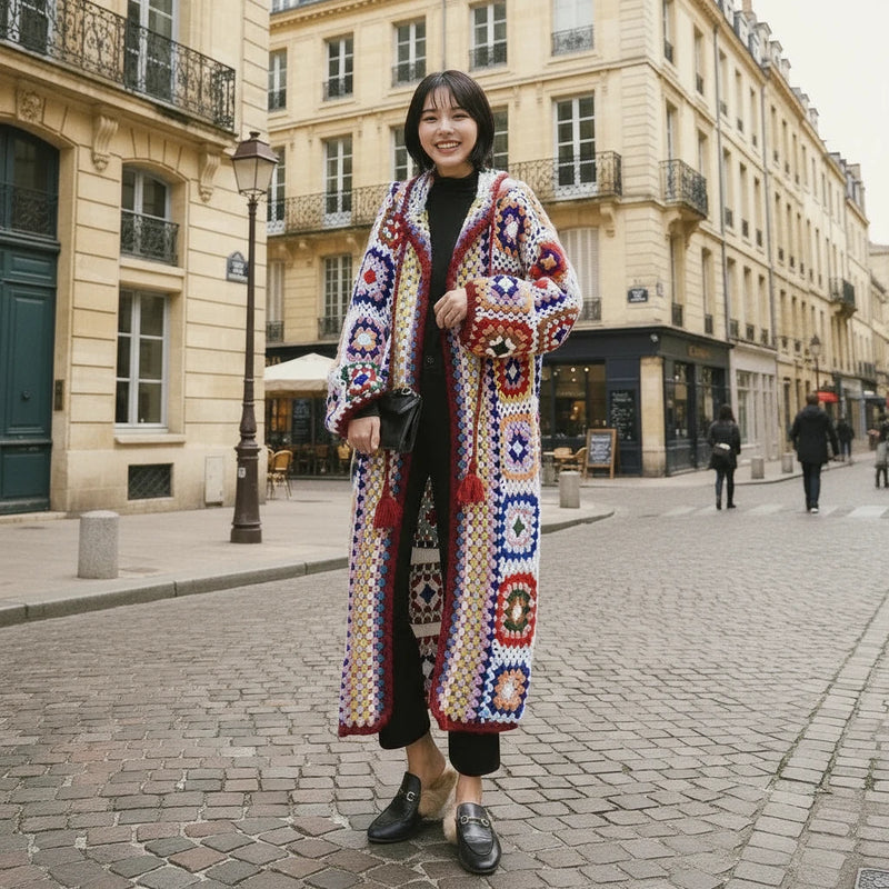 A woman standing against a wall, wearing a long, multicolored geometric patterned hooded crochet sweater coat.