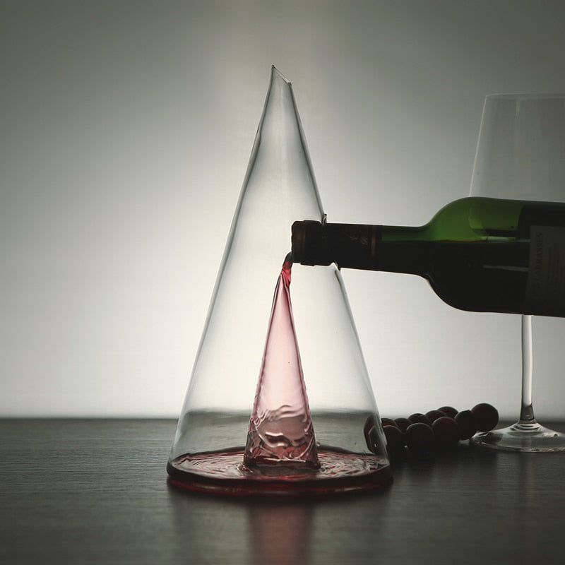 A transparent pyramid-shaped glass wine decanter with red wine being poured into it, set against a neutral background.