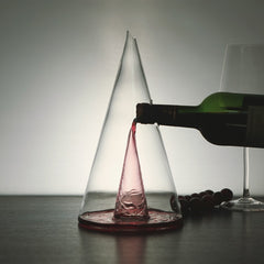 A transparent pyramid-shaped glass wine decanter with red wine being poured into it, set against a neutral background.