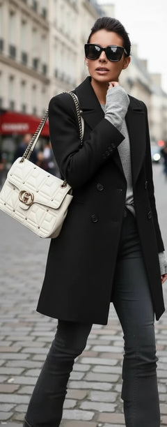 Woman wearing a black coat, gray sweater, and jeans with a white handbag.