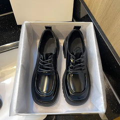 A pair of black thick sole lace-up leather shoes presented on a box with a hand holding one shoe.