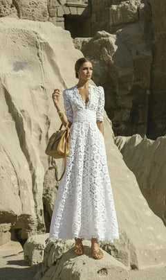 A woman wearing a white long lace dress with short sleeves, standing near a rocky beach edge.
