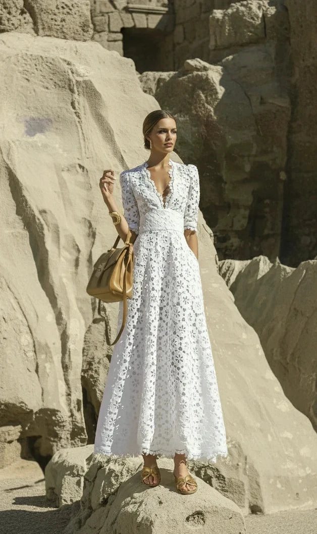 A woman wearing a white long lace dress with short sleeves, standing near a rocky beach edge.